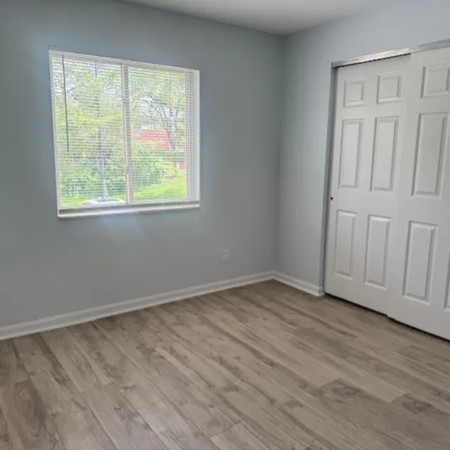 A bright, empty room with light blue walls and a window letting in natural light. The floor is made of light wood planks, and there is a double-door closet on the right side.