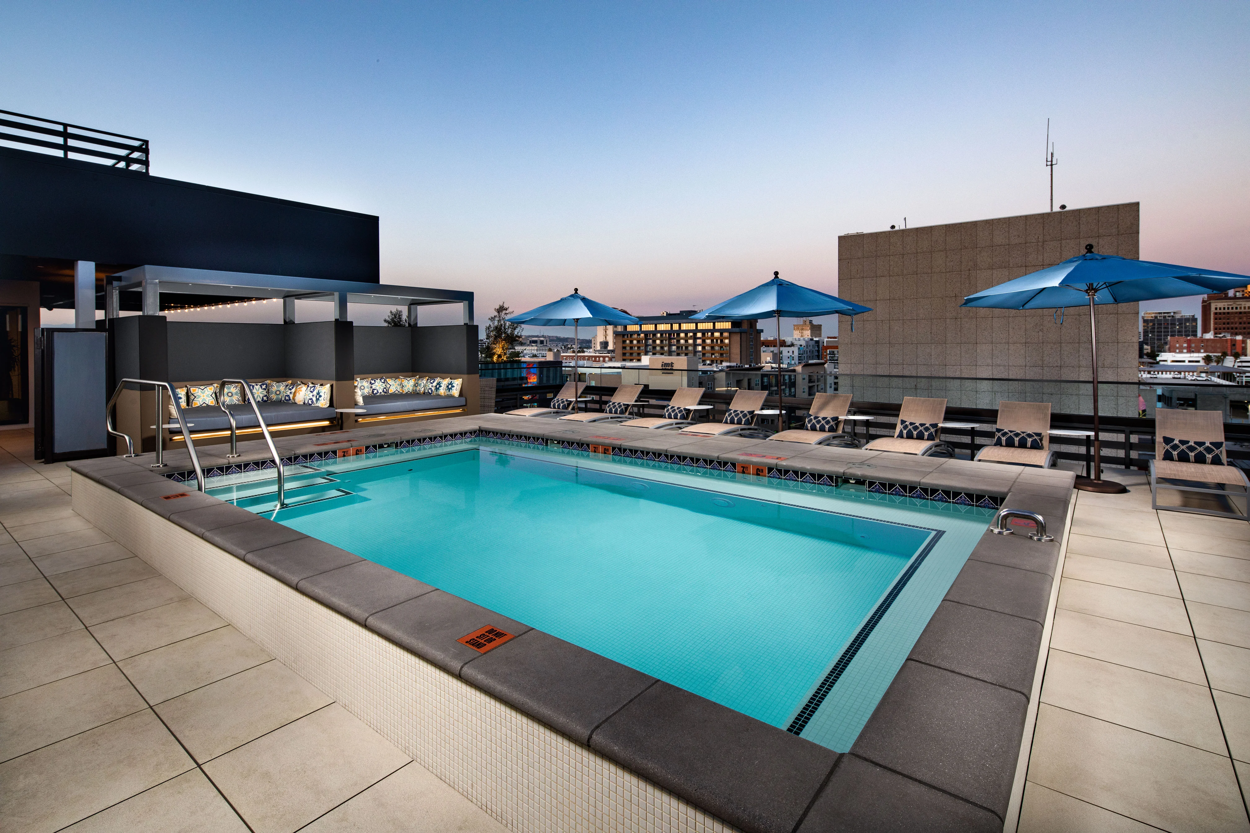 A rooftop pool with lounge chairs and blue umbrellas during sunset.