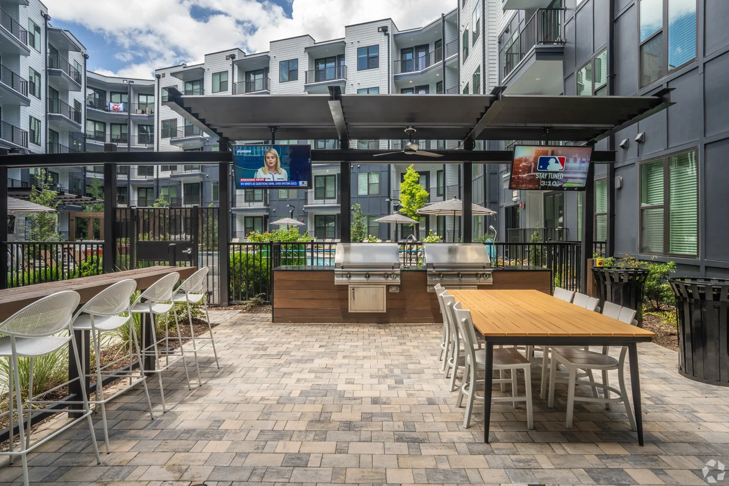 Outdoor Grilling Area in Apartment Complex A modern outdoor communal grilling area with a dining table, bar stools, and potted plants, surrounded by apartment buildings.