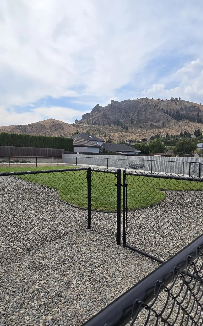A fenced dog park featuring a grassy area surrounded by gravel, with mountains in the background and a few buildings nearby.