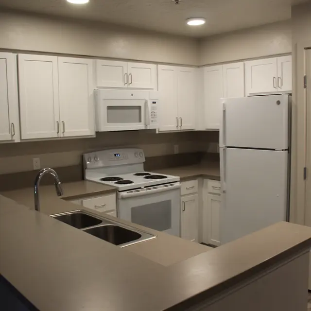 An interior view of a modern kitchen featuring white cabinetry, a microwave, a stove, and a refrigerator. The countertop is a neutral color with a double sink visible. Light fixtures are installed in the ceiling, illuminating the space.