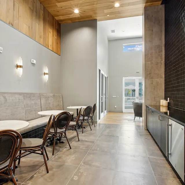 Modern Indoor Design with Seating and Beverage Station A modern indoor space showcasing a minimalist design with a wooden accent wall, plush seating area, round marble tables, and stylish chairs. Bright lighting enhances the atmosphere, giving it a welcoming feel. The opposite wall features a dark tile design with a beverage station.