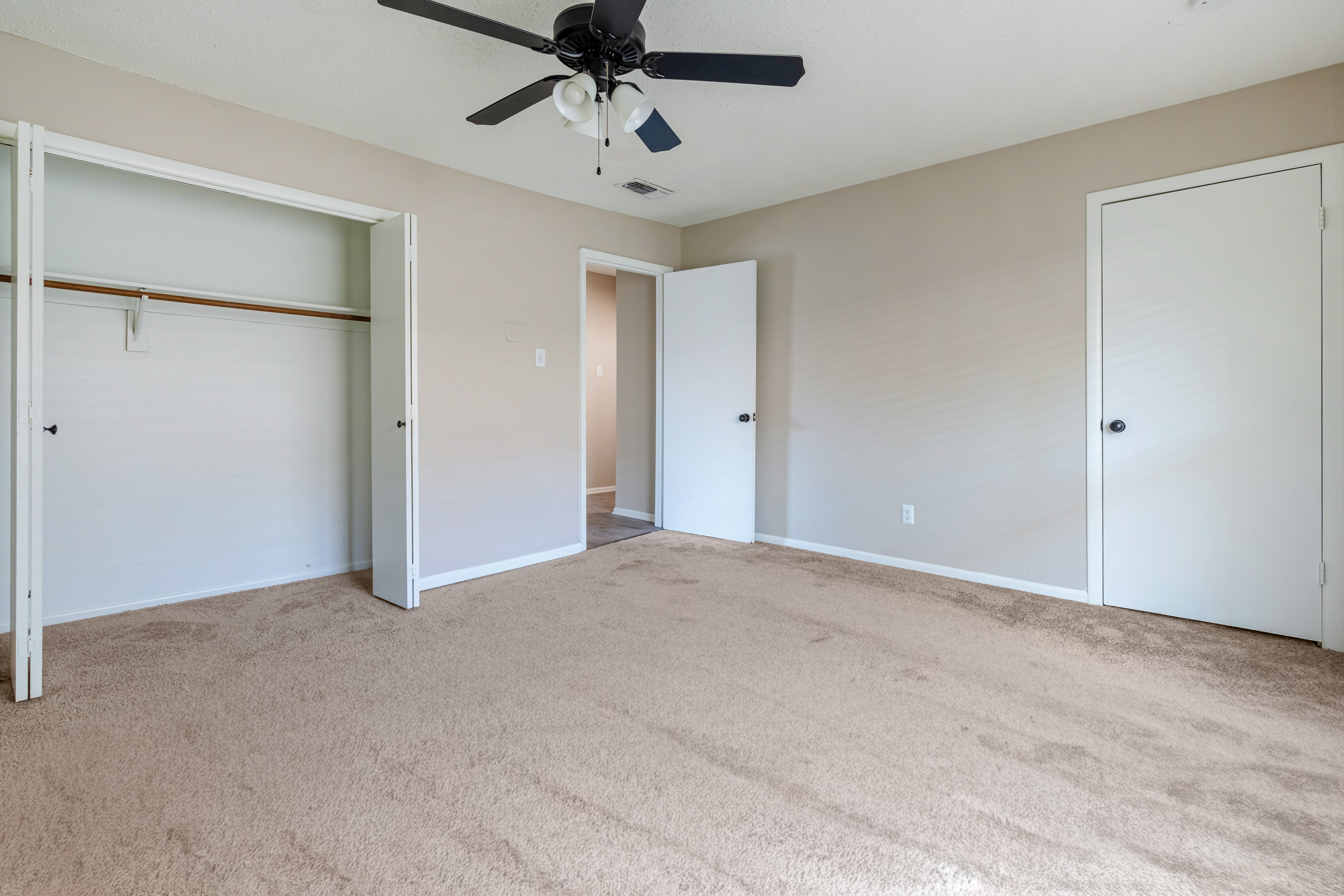 A vacant room featuring beige walls, carpeted flooring, and a ceiling fan. There are open doors to closets on the left and another door on the right leading to a different space. The room is well-lit with natural light.