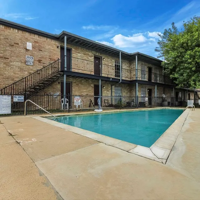 An outdoor swimming pool next to a two-story apartment building. The pool is clear and surrounded by concrete. The building features a brick facade with balconies. Chairs are placed near the pool, and trees can be seen in the background.