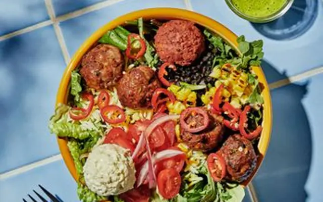 A vibrant salad bowl filled with mixed greens, meatballs, tomatoes, and sliced peppers accompanied by a small glass of green drink.