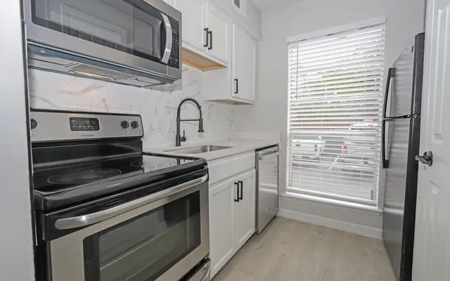 A modern kitchen featuring stainless steel appliances including an oven, microwave, and refrigerator. The kitchen has white cabinetry, a sink with a stylish faucet, and a window with blinds allowing natural light to enter.