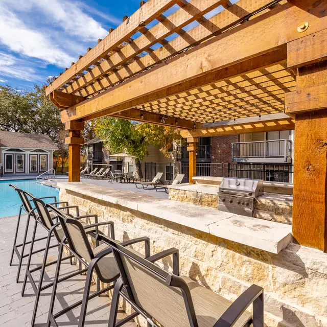 Inviting Poolside Retreat A swimming pool area featuring a stone bar under a wooden pergola with seating, surrounded by landscaped trees and an apartment building.