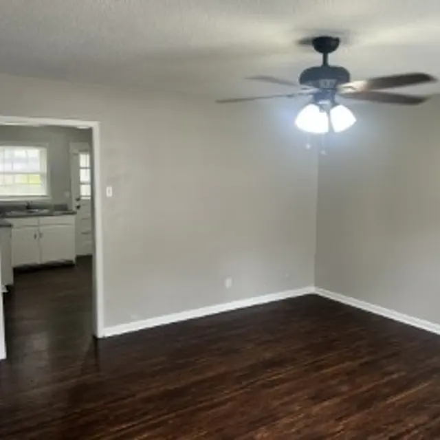 A spacious living room featuring dark wood flooring, a ceiling fan with three light bulbs, and beige walls. An entryway leads to a kitchen visible in the background.