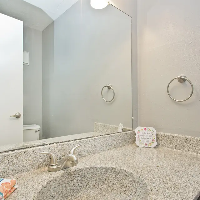 A clean, modern bathroom featuring a gray wall, a large mirror above a granite countertop, a sink with a faucet, and a towel ring. A door is visible to the left, leading to another area.