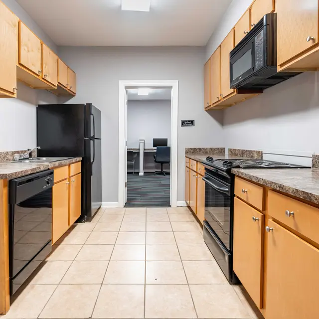 A spacious kitchen with wooden cabinets, black appliances including a refrigerator, dishwasher, and microwave, and a tiled floor. The entrance to another room is visible at the back.