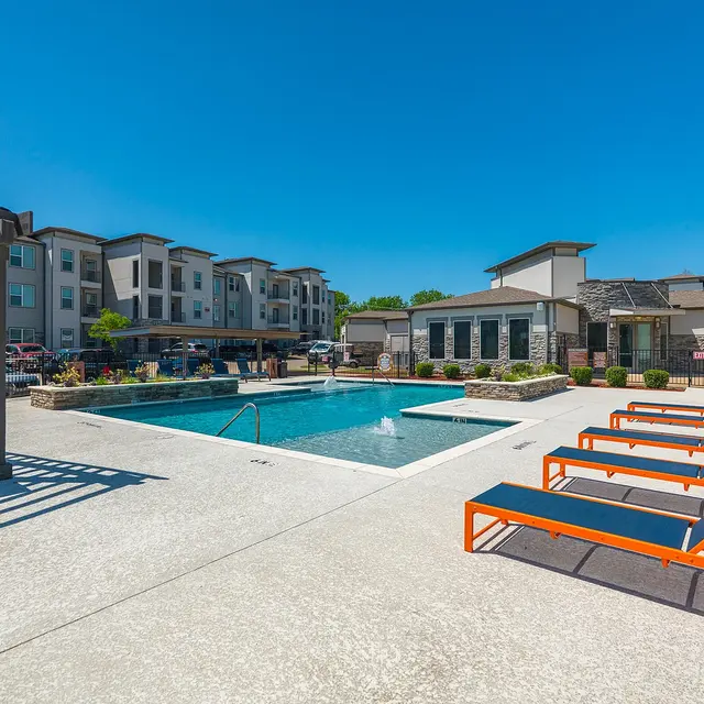 View of an apartment complex pool area featuring a swimming pool, lounge chairs, and surrounding buildings under a clear blue sky.
