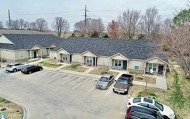 Aerial view of a residential housing complex featuring several single-story buildings with gray roofs, surrounded by a parking lot filled with cars. There are patches of grass and sparse trees in the vicinity, under a cloudy sky.