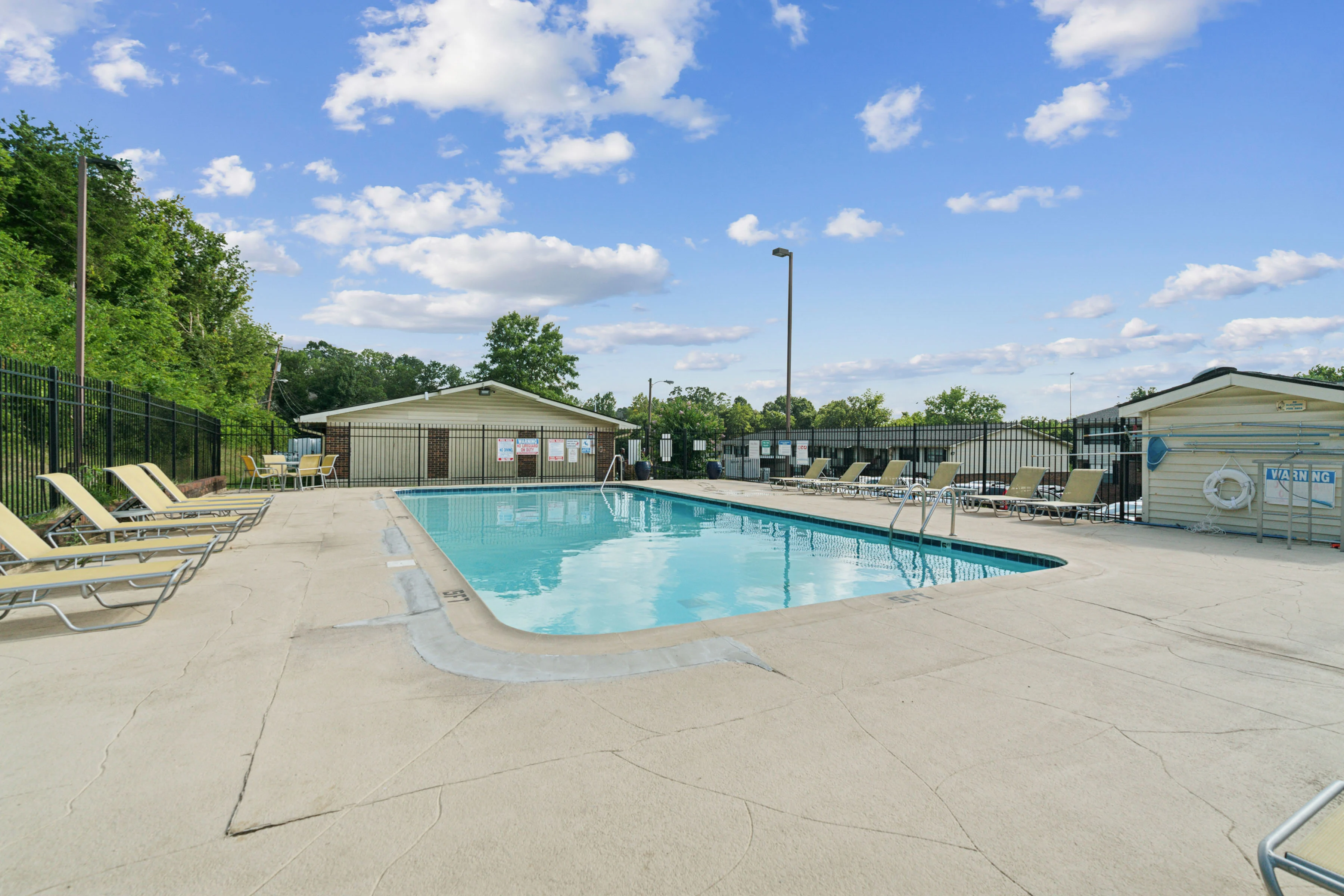 A clear blue swimming pool surrounded by lounge chairs and a fenced area under a bright, cloudy sky.