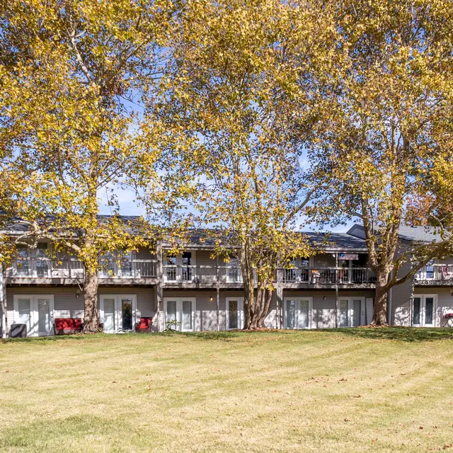 A modern hotel building surrounded by large, golden-leaved trees in autumn. The hotel features balconies and multiple doors facing a manicured lawn.