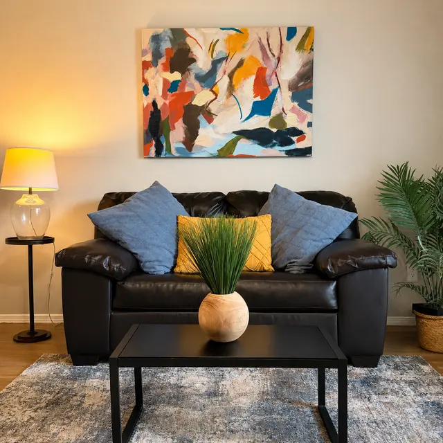 A cozy living room setup featuring a black leather couch with decorative blue and yellow pillows, a black coffee table with a round vase containing decorative grass, and a colorful abstract painting on the wall. A floor lamp is illuminated beside the couch, and there is a potted plant in the corner.