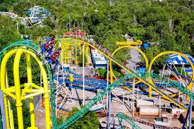 An aerial view of a vibrant amusement park featuring various roller coasters with tracks in green, yellow, and blue colors, surrounded by lush greenery.