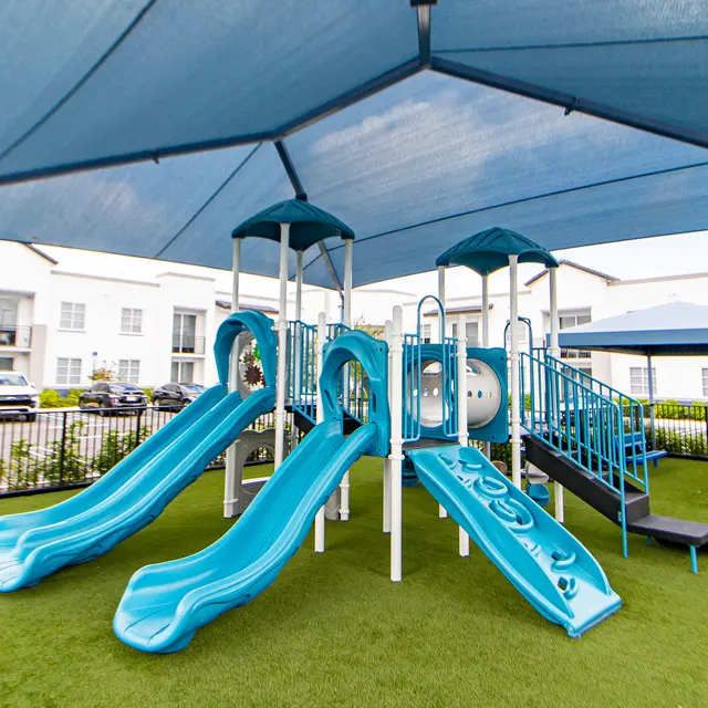 Blue Playground Slides Under Shade A playground featuring blue slides and a shaded area with a tent. The ground is covered in green artificial grass. There are several structures including a climbing area and play panels, set in a residential community environment.