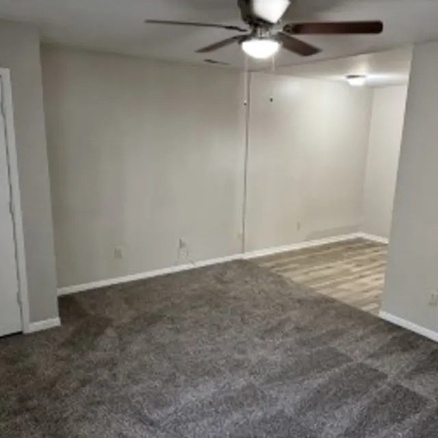 An empty room with light brown carpet and light gray walls. A ceiling fan is visible, and there is a white door on the left. A transition to another room is visible at the right side.