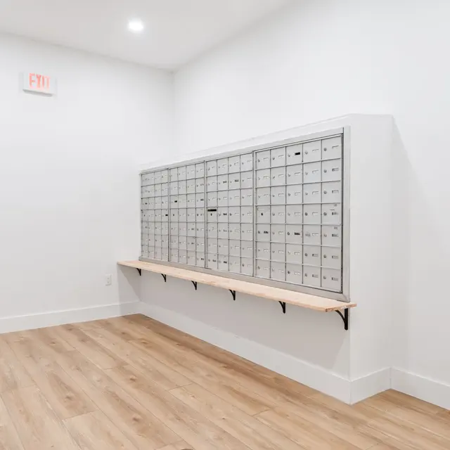 A modern mailbox area with individual mail slots, mounted on a wall. The setting features a minimalist design with light wood flooring and white walls, under bright lighting.