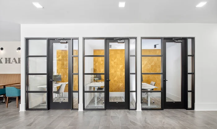 Contemporary Office Meeting Rooms A modern office environment featuring several glass-doored meeting rooms, with a visible yellow accent wall in the background and two tables and chairs visible inside the rooms.