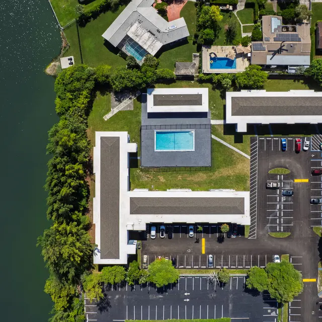 Aerial view of an apartment complex with a swimming pool surrounded by green areas and parking spaces.