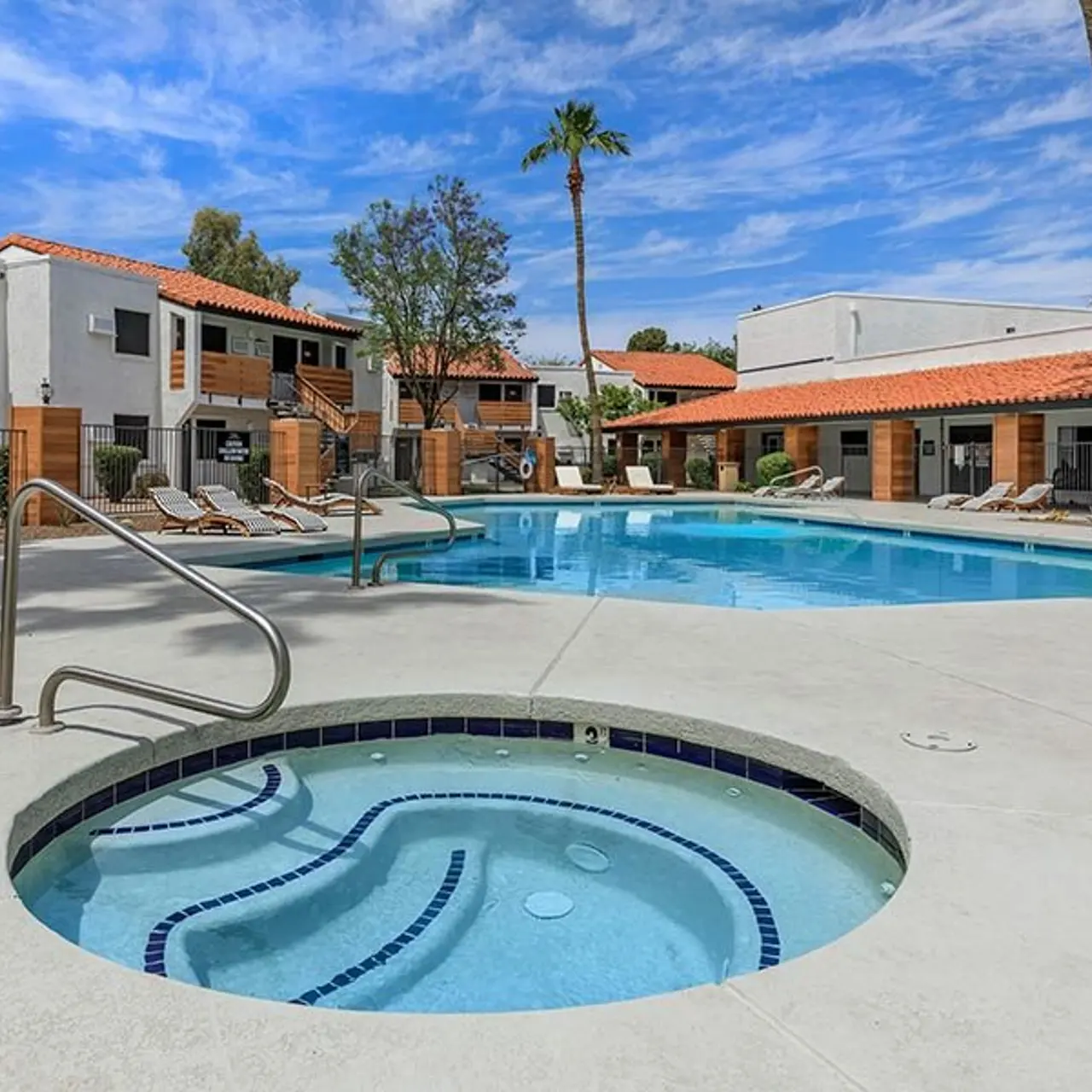 A view of a swimming pool area featuring a larger pool and a hot tub, surrounded by lounge chairs and nicely landscaped greenery, with a bright blue sky in the background.