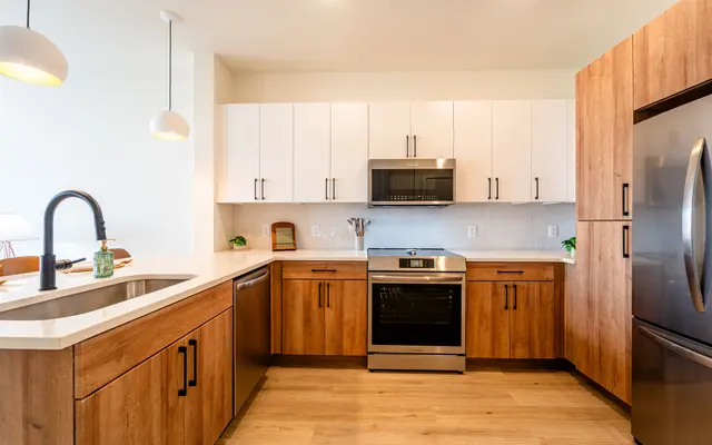 A modern kitchen featuring a combination of wooden and white cabinetry, stainless steel appliances including an oven and microwave, and a spacious countertop with a sink. The flooring is light wood, and there are pendant lights hanging above the counter.