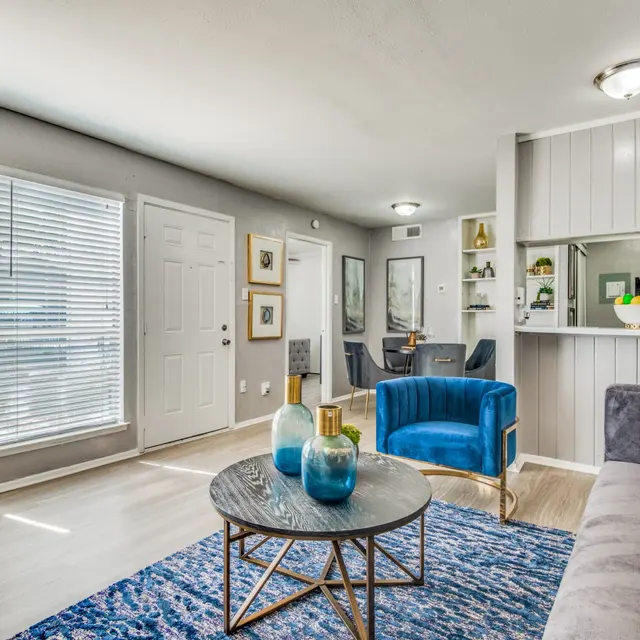 A contemporary living room featuring a grey sofa, a round coffee table with decorative items, and blue armchair. There are large windows with blinds allowing natural light to enter, and a dining area visible in the background.