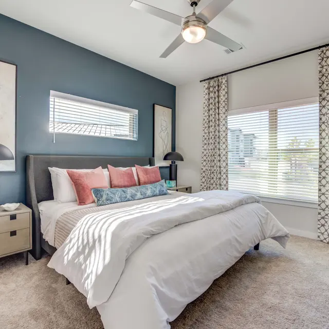 A modern bedroom featuring a bed with a grey headboard, decorative pillows in shades of pink and blue, a nightstand with a lamp, and a window with patterned curtains.