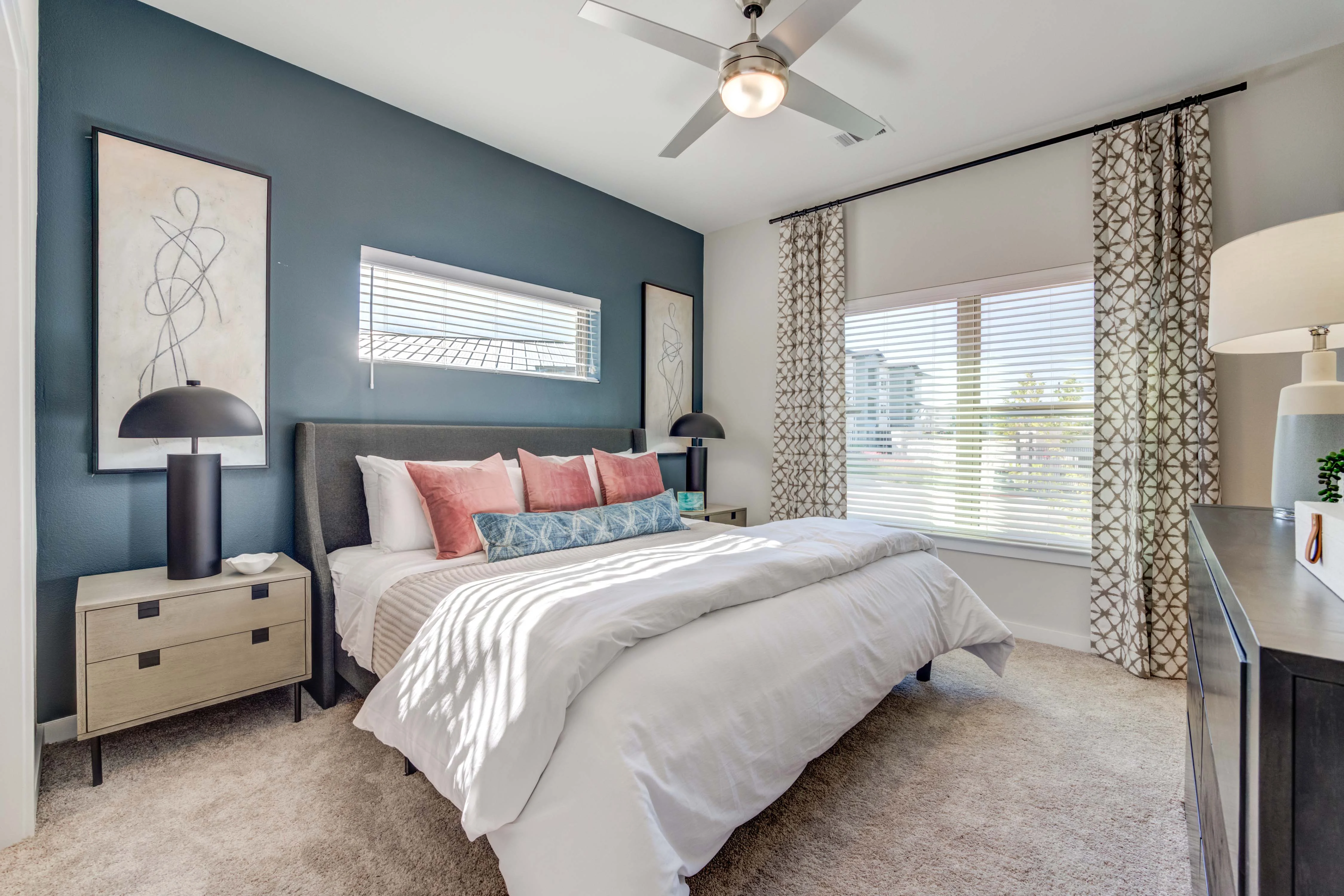 A modern bedroom featuring a bed with a grey headboard, decorative pillows in shades of pink and blue, a nightstand with a lamp, and a window with patterned curtains.