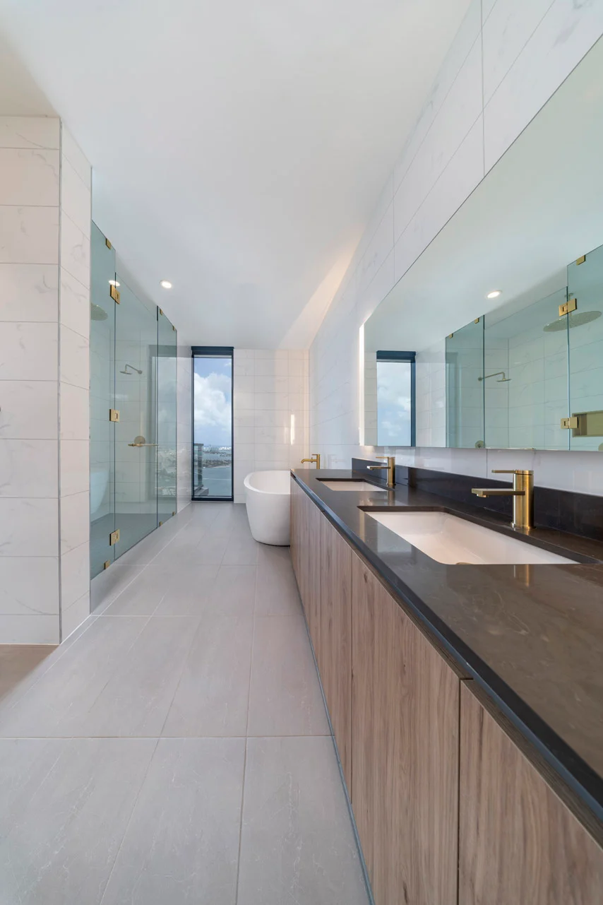 A spacious modern bathroom featuring a large wooden vanity with a black countertop, two sinks, and gold fixtures. A sleek white freestanding bathtub is situated next to a glass shower with gold accents. The walls are adorned with white tiles, and large mirrors enhance the airy feel of the space. Natural light floods in from a window.