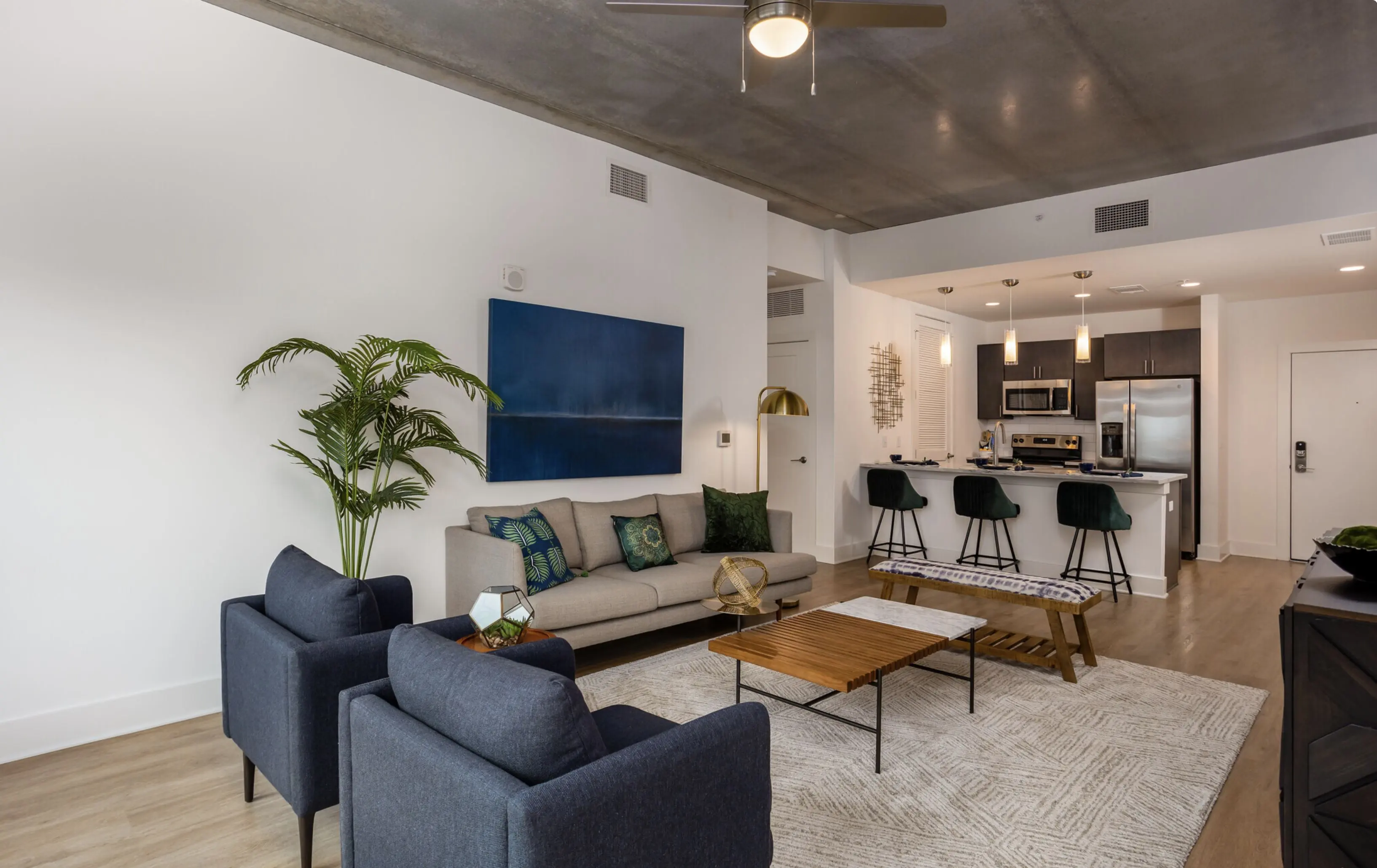 A stylish and modern apartment living room featuring a cozy seating area with gray sofas, a coffee table, and a decorative plant. The room has a sleek design with a concrete ceiling and an open kitchen in the background.