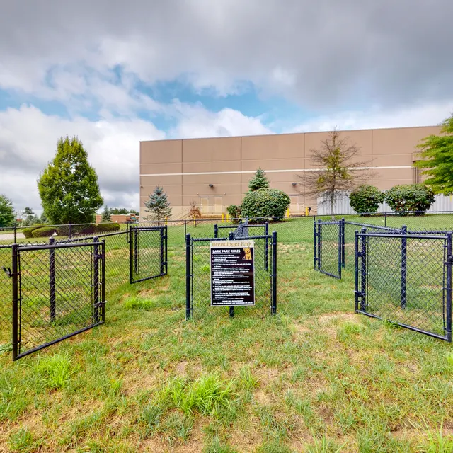 Dog Park Area An open area of a dog park enclosed by a chain-link fence, featuring several gates and green grass under a cloudy sky.