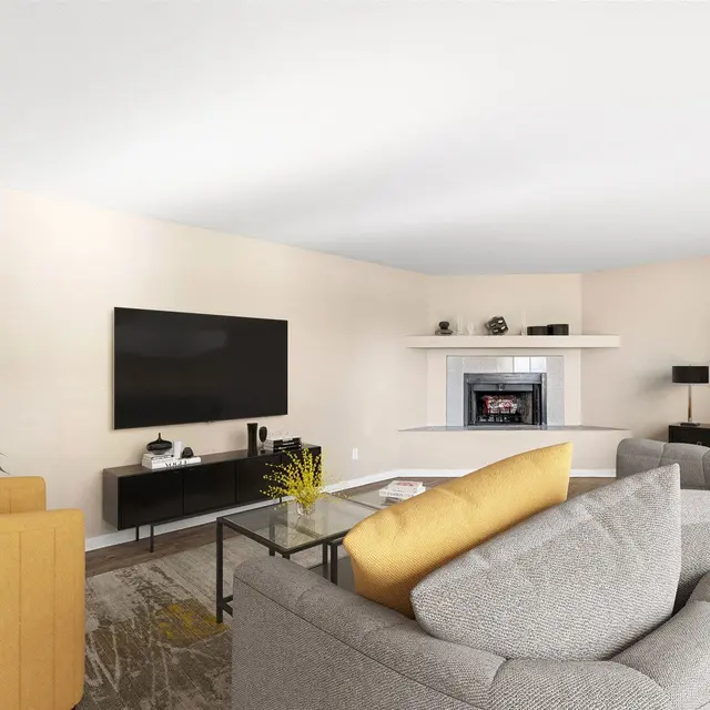 A modern living room featuring a gray couch, a yellow accent chair, a coffee table, and a flat-screen TV mounted on the wall. There is a decorative plant in the corner and a simple fireplace with a mantel in the background. The walls are painted a soft beige color, and a piece of abstract artwork is displayed on the wall.