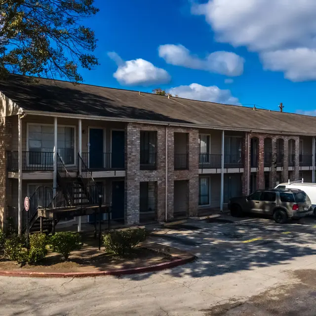 A two-story apartment building with a mix of brick and siding, featuring several balconies and parked cars in front.