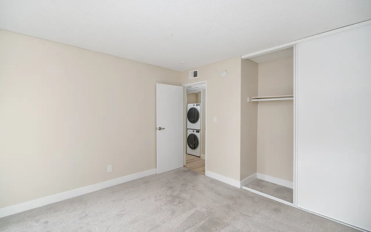 A spacious, empty bedroom with beige walls and carpeted flooring. There is a white closet with sliding doors to the right. A door leads to a small laundry area with stacked washing machines visible through an open door.