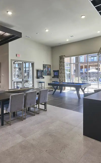 A modern lounge area featuring a dining table with chairs, a billiards table, a large window with natural light, and a feature wall with a television.