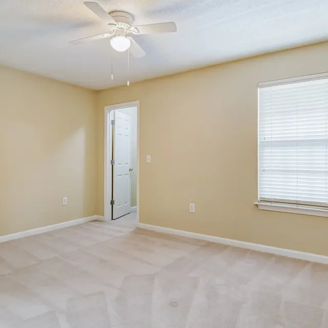 An empty room with beige walls and light beige carpeting. A ceiling fan is visible, and there's a window with white blinds on the right. A door leads to another area, and the space is well-lit.