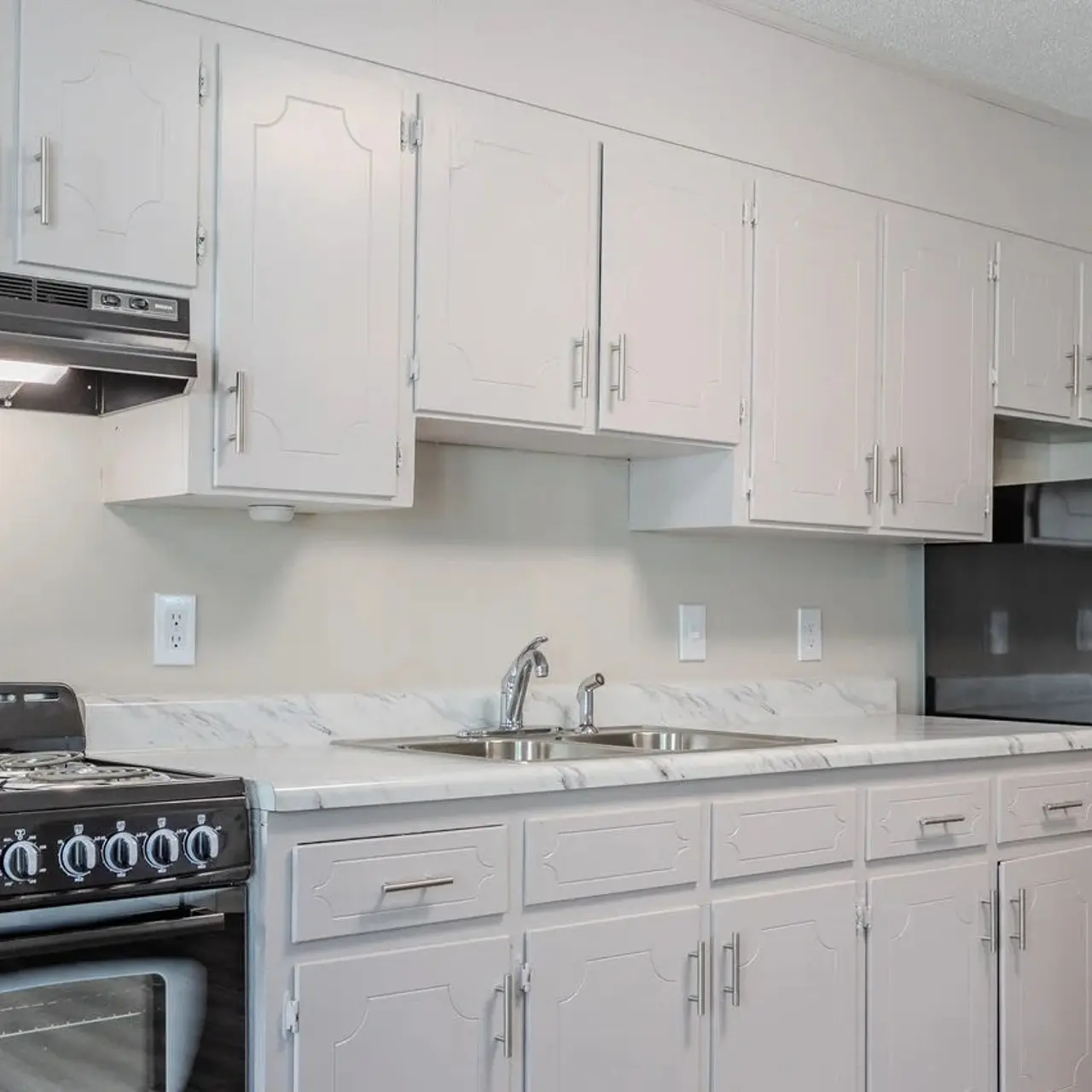 A modern kitchen featuring white cabinets, a black stove with a hood, a stainless steel refrigerator, and a sink with a faucet, all set against a light-colored wall.