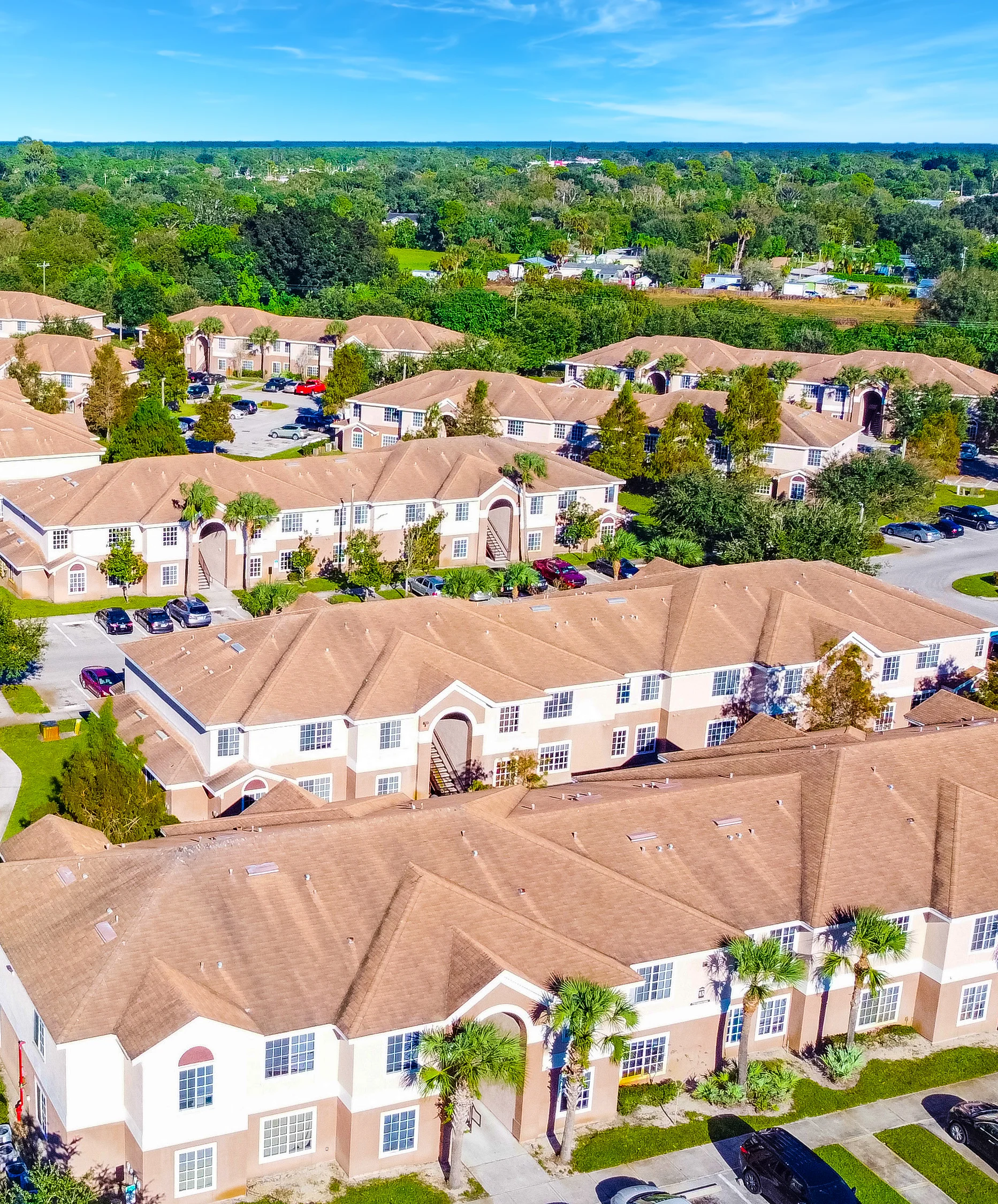Aerial View of Apartment Complex Aerial view of an apartment complex featuring multiple buildings with brown roofs surrounded by lush greenery and parked cars.