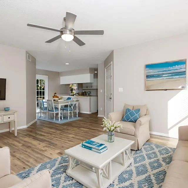 Bright and airy living room featuring a beige sectional sofa and a blue accent chair. A coffee table sits on a patterned rug, with a flat-screen TV mounted on the wall. The room leads into a dining area with a modern kitchen visible in the background.