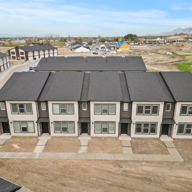 Aerial view of newly constructed townhomes with a gray roof and modern design. Sidewalks lead up to each unit, and the surrounding area is mostly bare land with some vegetation.