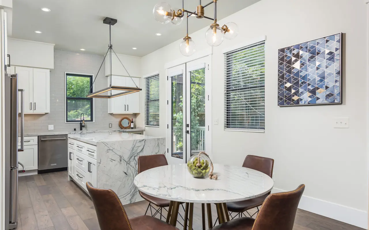 A modern kitchen and dining area featuring a round marble table with brown chairs, a white kitchen with marble countertops, and large windows letting in natural light.