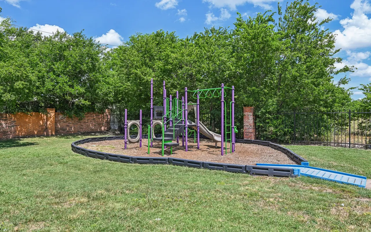 A playground structure with purple and green colors surrounded by grass and trees under a blue sky with fluffy clouds.