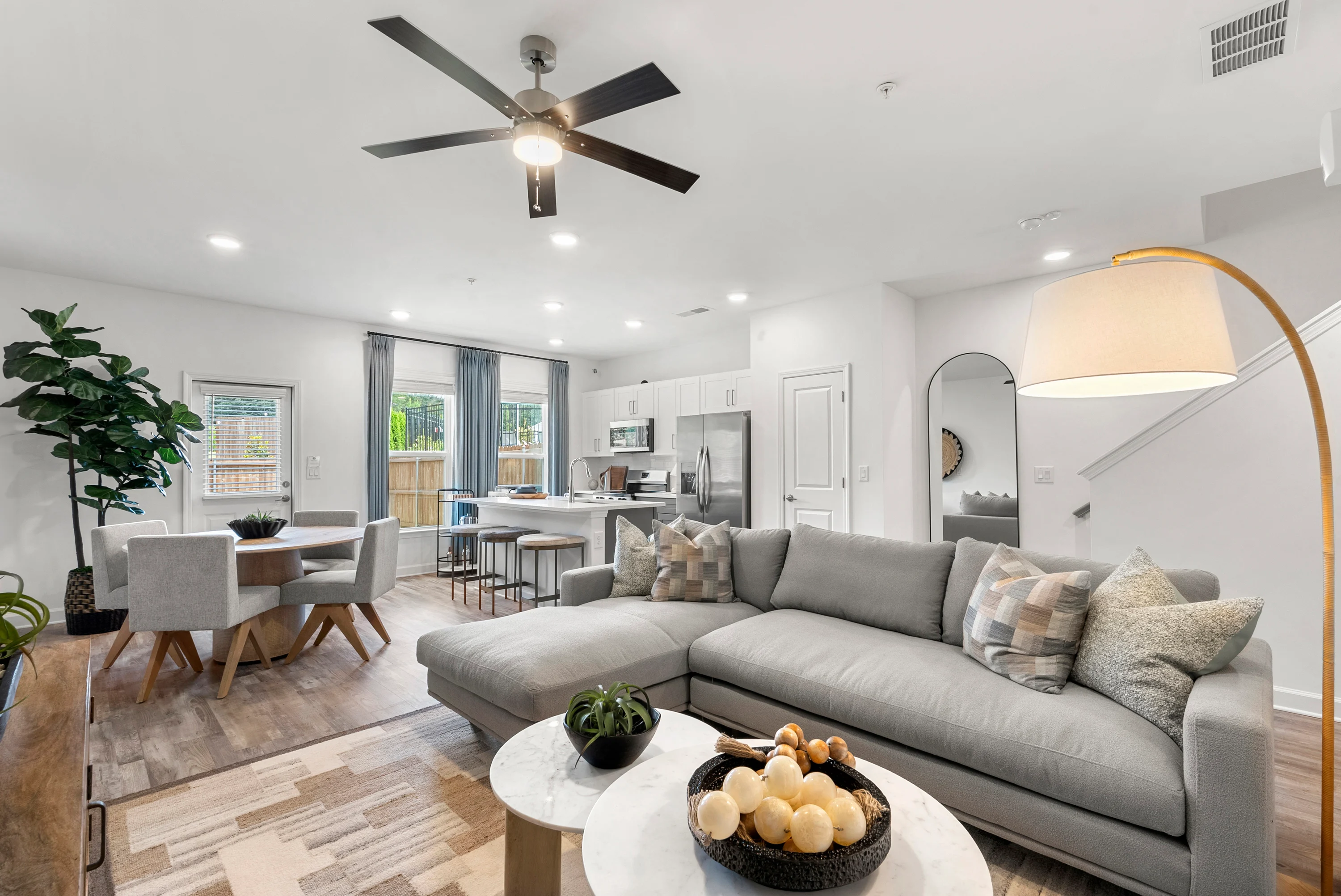 Contemporary Living and Dining Area A modern living room featuring a gray sectional sofa, a round coffee table with decorative items, and a dining area with a table and chairs. A potted plant adds greenery to the space, and a fan is mounted on the ceiling.
