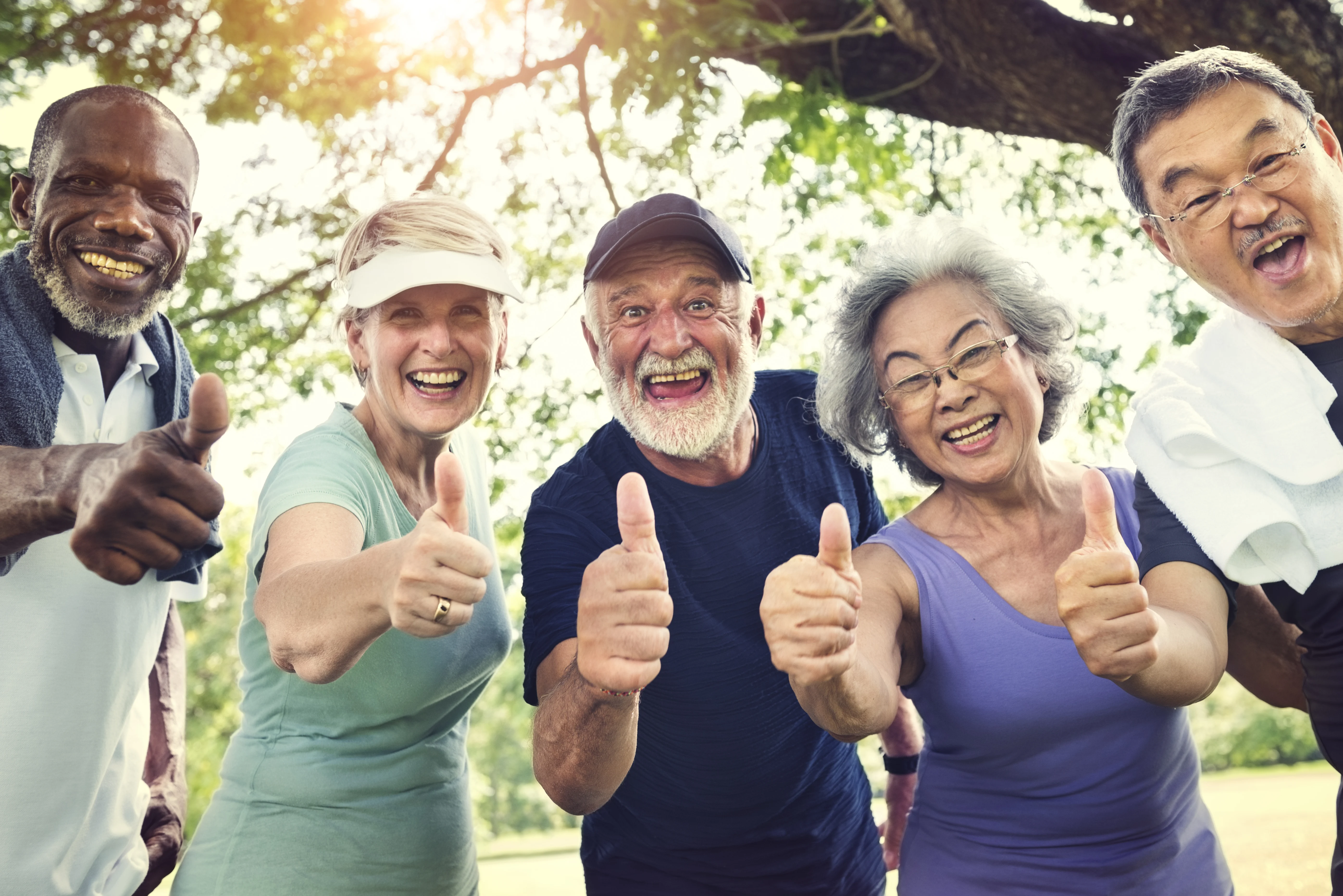 A diverse group of five senior adults standing outdoors, smiling and giving thumbs up. They exude positivity and joy, with trees and bright sunlight in the background.