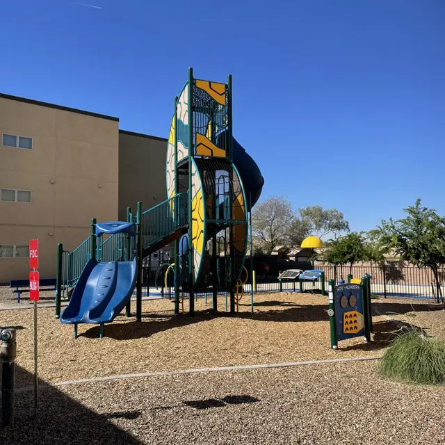 A colorful playground with a large slide, play structures, and a sunny blue sky in the background. The area is surrounded by a fence and buildings.