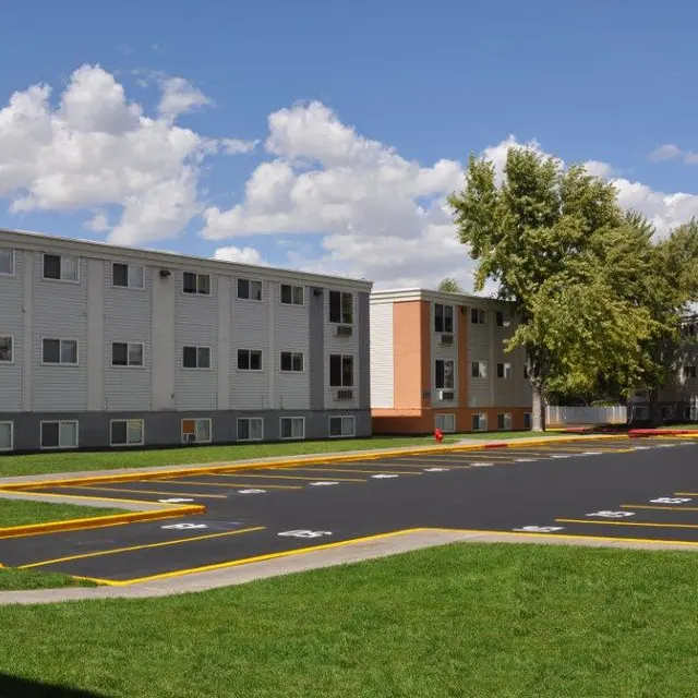 A modern apartment complex with multiple buildings and a parking lot surrounded by grass and trees under a partly cloudy sky.