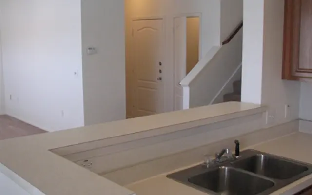 Open Kitchen and Living Area An open kitchen area viewed from the perspective of the countertop. The kitchen has a double sink and wooden cabinets. In the background, a staircase is visible leading to a second floor, along with a door and hallway.