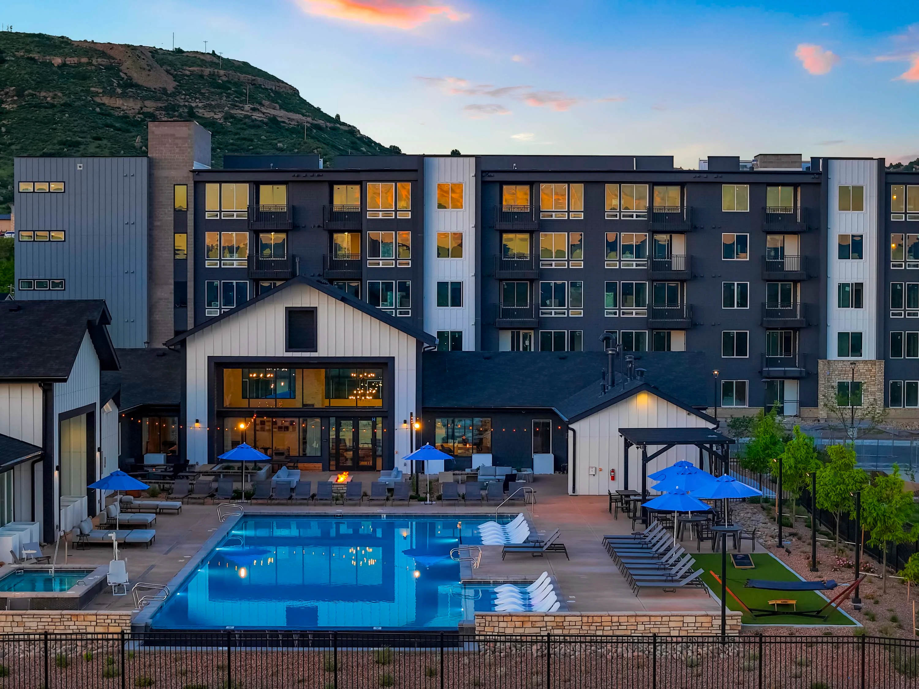 View of a modern apartment complex featuring a swimming pool and outdoor lounge area, with mountains in the background during sunset.